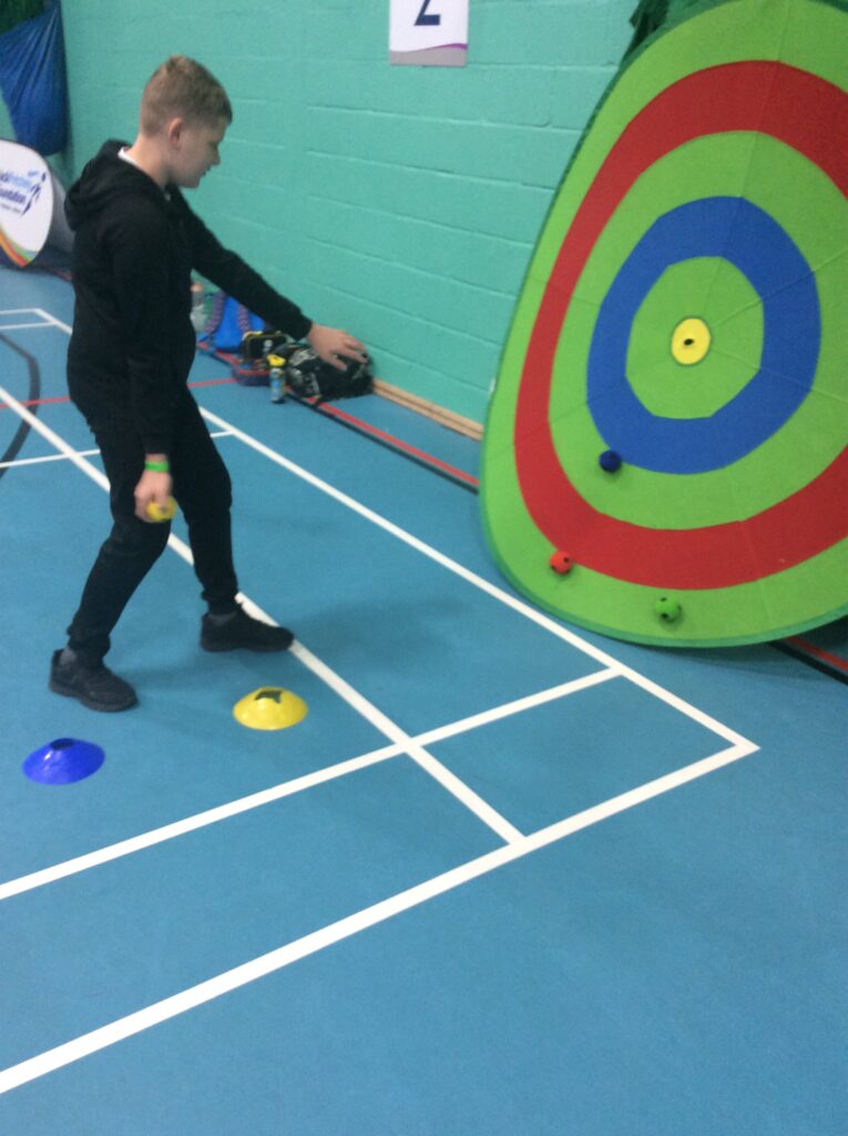 A young, male student can be seen having just thrown a dart at a large plastic target in the Sports Hall, as part of a school-wide competition.