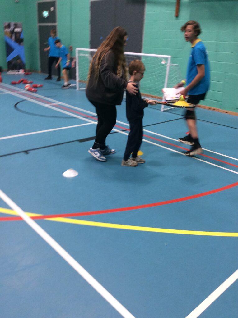 A young, male student is pictured being assisted in a sporting activity in the Sports Hall by a female member of staff.
