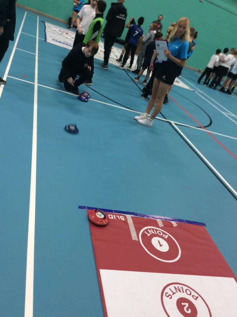 A male student is pictured kneeling down in the Sports Hall, having just thrown a puck towards a target on the floor. An older female student is seen standing beside him, recording his score.