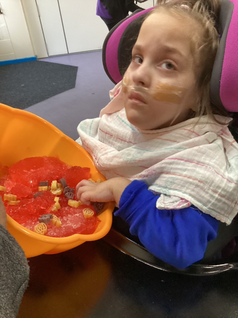 A young girl in a wheelchair is pictured looking towards the camera, whilst holding an orange bucket in her hands with toys inside.
