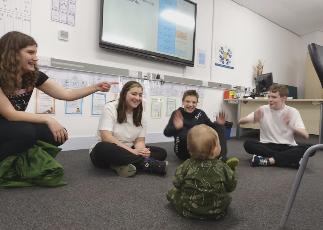 A small group of students are pictured sat on the floor together in the middle of a classroom, smiling and interacting with a toddler who is sat with them.