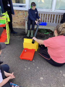 A female member of staff can be seen supervising a young male student with an activity involving plastic tubs and tubes outdoors.