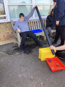 A member of staff can be seen supervising a young male student with an activity involving plastic tubs and tubes outdoors.