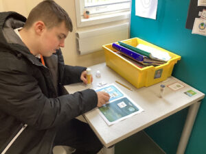 A male student is pictured sat at his desk indoors, using a glue stick to add some images to a sheet of paper in front of him.