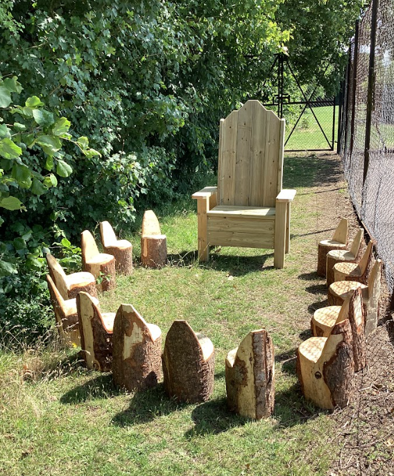 This picture shows an outdoor setting with a unique seating arrangement. In the center of a grassy area, there's a large, grand-looking wooden chair, resembling a throne, made of light-colored wood. Arranged in a circle around this central "throne" are several smaller, rustic seats. These seats appear to be made from tree stumps or logs, with a cut-out section to form a backrest, giving them a natural and simple look. There are at least eleven of these smaller seats visible, creating a gathering space. The area is surrounded by lush green foliage, suggesting it's in a natural or semi-natural environment like a garden, park, or woodland edge. To the right, a chain-link fence is visible, and beyond that, there appears to be more greenery and possibly some structures in the distance. The overall impression is that of an outdoor storytelling circle or a designated area for imaginative play, perhaps a "fairy circle" or "nature's meeting place."
