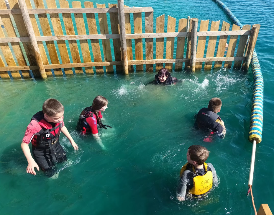 In the image, several people are in the water, which appears to be part of an outdoor water activity or park. There's a wooden barrier or fence extending into the water, creating an enclosed area. Some individuals are wearing wetsuits and life vests, suggesting they are participating in a water sport or recreation. One person in the back appears to be splashing or playing in the water, creating ripples. The overall scene conveys a sense of leisure and enjoyment in an aquatic setting.
