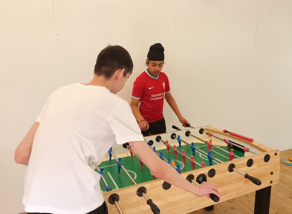 The image shows two young people engaged in a game of foosball (table football). The boy on the left, viewed from behind, is wearing a white t-shirt and is actively playing, with his hands on the game rods. The boy on the right is wearing a red Liverpool FC jersey and is also focused on the game. The foosball table is made of wood with green playing surface and traditional red and blue player figures. They are indoors, in a room with plain white walls and a light wooden floor.