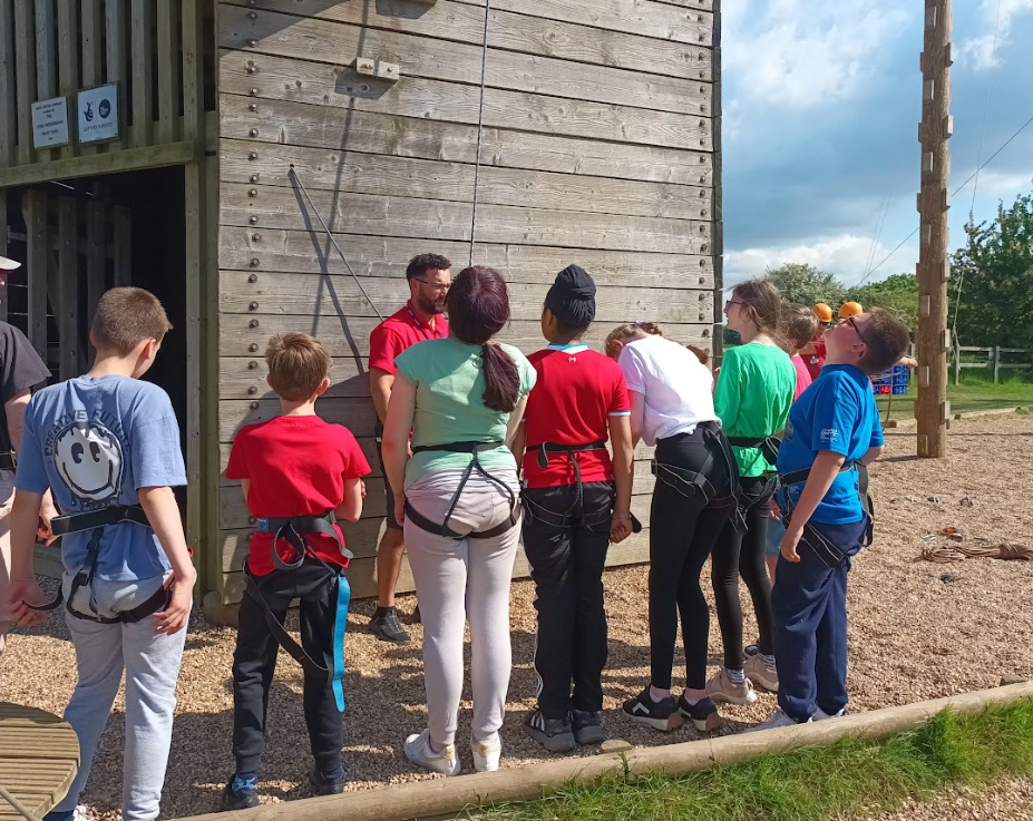 The image shows a group of young people gathered around a large wooden structure, which appears to be part of a high ropes course or climbing wall. Most of the young people are wearing harnesses, indicating they are about to participate in an aerial activity. An adult, possibly an instructor, is standing in the middle, addressing the group. The ground is covered with wood chips, a common safety surfacing for such activities. In the background, there's another tall wooden pole structure, and the sky is partly cloudy. This scene suggests a group activity at an outdoor adventure center.