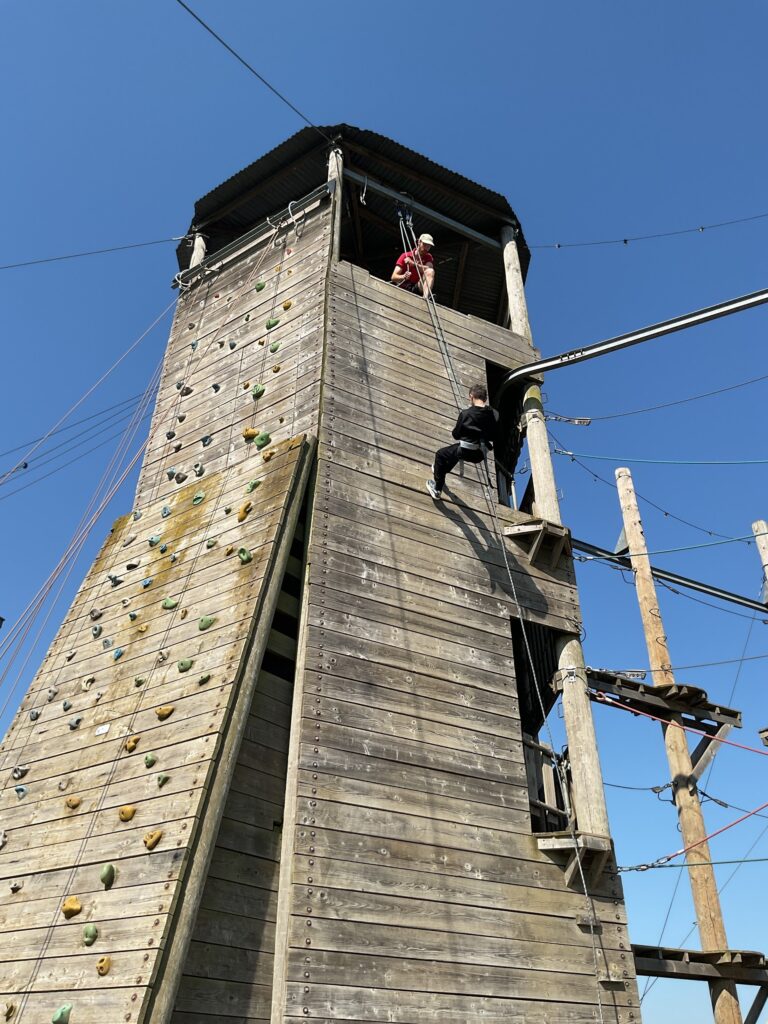 The image displays a tall, wooden adventure tower under a clear blue sky. On the left side of the tower, there's a climbing wall with various handholds. On the right side, a person in dark clothing is actively abseiling down the side of the tower, controlled by a rope. Another person is visible at the top of the tower, likely assisting or supervising the abseiler. There are also ropes and cables extending from the tower, suggesting it's part of a larger high ropes or adventure course. This scene shows an abseiling activity in progress at an outdoor adventure center.