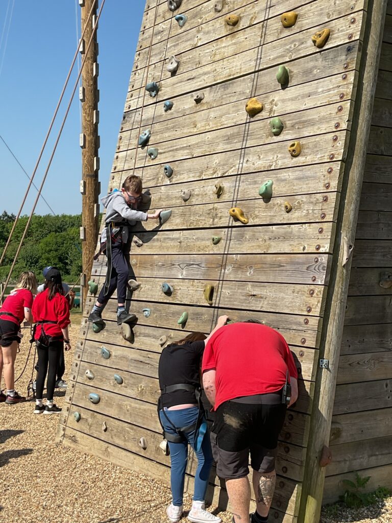 The image shows an outdoor climbing activity on a wooden climbing wall. A young person, wearing a grey hoodie and harness, is actively climbing the wall, reaching for a handhold. Below, several other individuals, some also wearing harnesses and red tops, are gathered around the base of the wall, seemingly either waiting their turn, belaying, or receiving instructions. The ground is covered with gravel or wood chips. In the background, there's a wooden pole with ropes, and green trees under a clear blue sky, indicating a sunny outdoor setting.