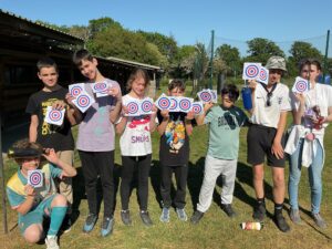 The image shows a group of about nine young people standing outdoors on a grassy area under a clear blue sky. Each of them is holding up one or more paper targets, all of which feature red, white, and blue bullseye patterns, suggesting they have just participated in a shooting or archery activity. They appear to be proud of their targets, with some smiling at the camera. The background consists of trees, a building with a green roof, and a net, possibly from another sports area. They are dressed in casual clothing, and the overall mood is one of accomplishment and enjoyment after an activity.