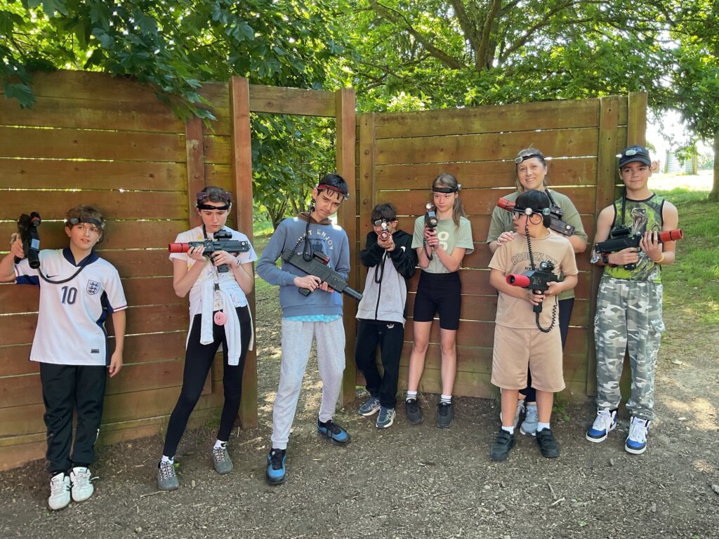 The image displays a group of eight people, predominantly young individuals, posing outdoors with laser tag equipment. They are standing in front of a wooden fence or structure, with trees and foliage in the background, suggesting a natural or wooded area for their game. Each person is holding a laser tag gun and wearing a headband with sensors, typical gear for this activity. They are dressed in casual clothing. The person in the middle-right appears to be an adult, possibly a supervisor or instructor. Everyone is looking towards the camera, and the atmosphere seems to be one of readiness and excitement for the game.