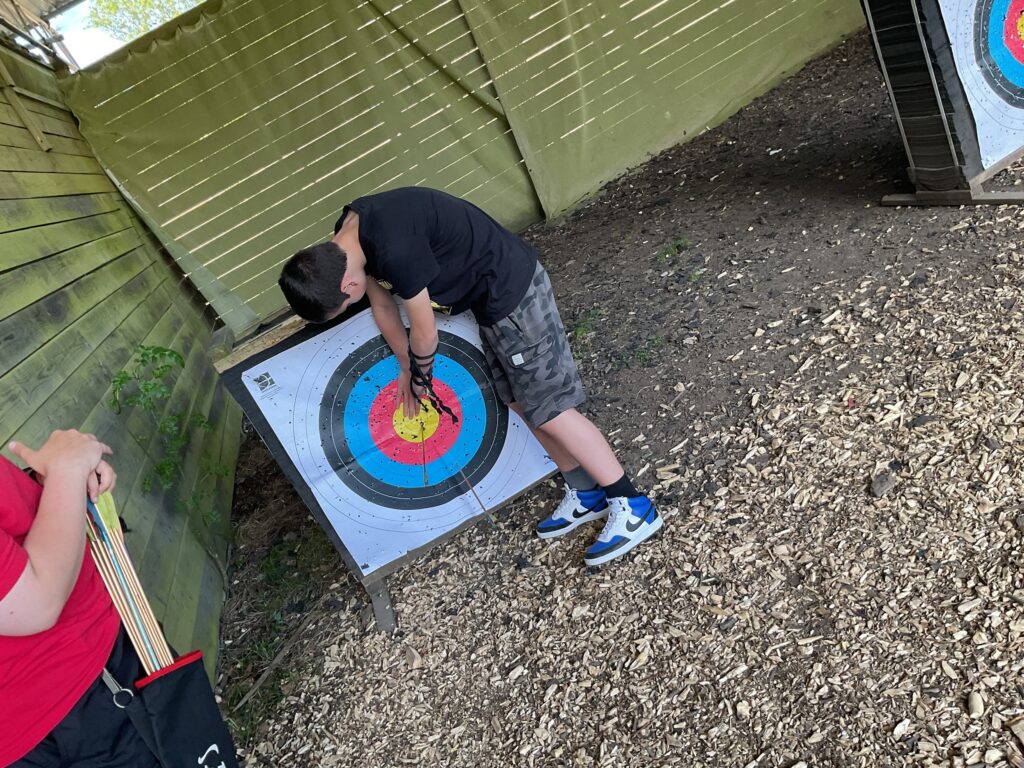 The image shows a person retrieving arrows from an archery target. The target is a standard multi-colored bullseye. The ground is covered with wood chips, suggesting an outdoor or semi-outdoor archery range. Another person's arm is visible on the left, holding a quiver full of arrows. In the background, there's a wooden fence or structure, and another archery target can be partially seen on the right. This scene depicts an archery activity.