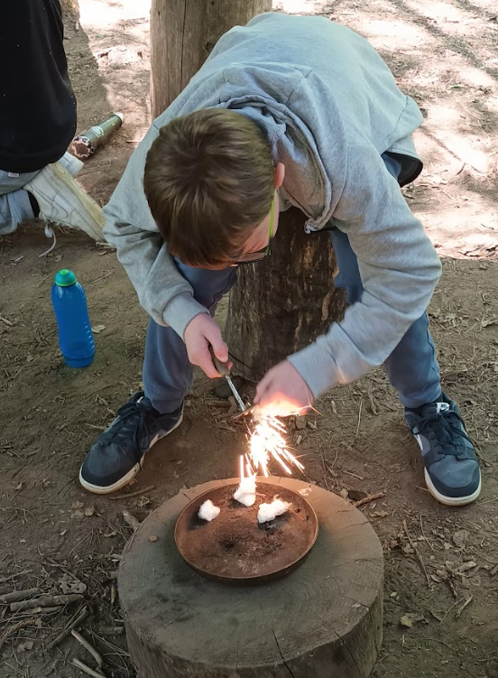 The image shows a young person actively engaged in a fire-starting activity outdoors. They are leaning over a metal plate, possibly a fire pan, which is placed on a tree stump. The person is using a ferro rod or similar fire starter to create sparks, which are igniting small pieces of tinder (likely cotton wool) on the plate, causing them to flare up. The setting appears to be a wooded area, with dirt and tree stumps on the ground, suggesting a survival skills or outdoor education program.