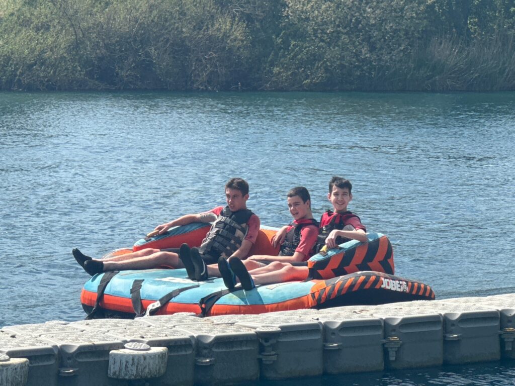 The image shows three young individuals sitting on an inflatable towable tube in a body of water, likely a lake or large pond. They are all wearing life vests, indicating they are participating in a water activity that involves being on the water. The inflatable device has "JOBE" visible on its side, a brand known for watersports equipment. They appear to be relaxed and enjoying the sunny weather. In the background, there are trees and foliage along the water's edge. They seem to be near a floating dock or platform.