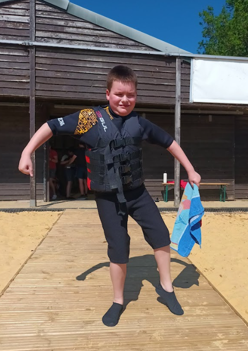 In the image, a young boy is standing on a wooden walkway, posing with his arms out and one leg slightly bent. He is dressed for water activities, wearing a black short-sleeved wetsuit with yellow patterns on the chest (the brand "GUL" is visible), a black life vest with multiple straps, and black water shoes. He is holding a blue and orange towel in his left hand. The background features a wooden building, possibly a changing room or activity center, and there's sand around the walkway. The bright sunlight suggests it's a clear, sunny day. The boy appears to be ready for or just finished with a water activity.