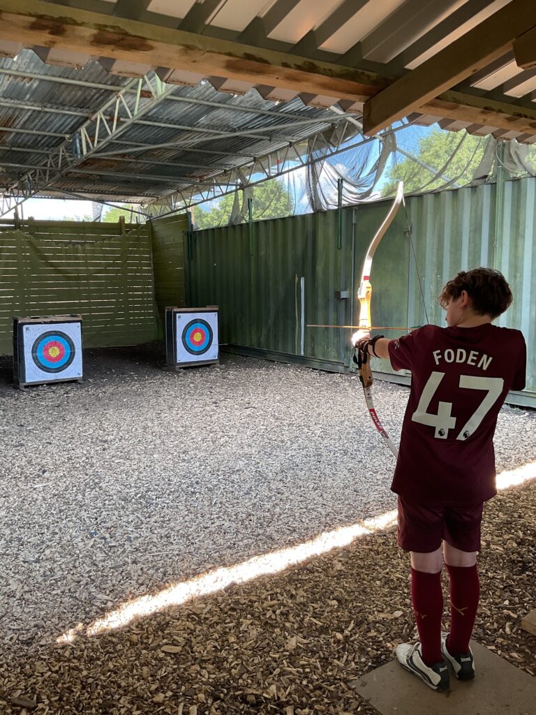The image captures an archery session in what appears to be a covered outdoor range. A young person, viewed from behind, is holding a bow and arrow, aiming at one of two archery targets set up in front of them. The person is wearing a maroon sports jersey with "FODEN" and the number "47" on the back, and matching shorts and socks. The ground is covered with wood chips. The range has a corrugated metal roof and walls, suggesting a dedicated area for the activity. Trees and blue sky are visible beyond the range structure, indicating an outdoor setting.