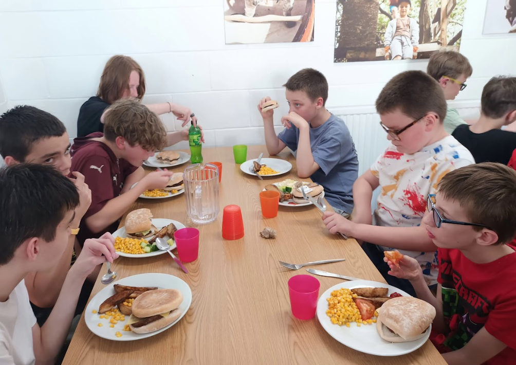 The image shows a group of young people, likely students, sitting around a long wooden table, eating a meal together. Each person has a plate with food that appears to include burgers or sandwiches, corn, and possibly potatoes or chips. There are also drinks in plastic cups and a large jug of water in the center of the table. They are dressed in casual clothes. The setting appears to be a cafeteria or dining hall, possibly at a school or a camp, with pictures on the white walls in the background. They are actively engaged in eating and some are looking at each other or at their food.