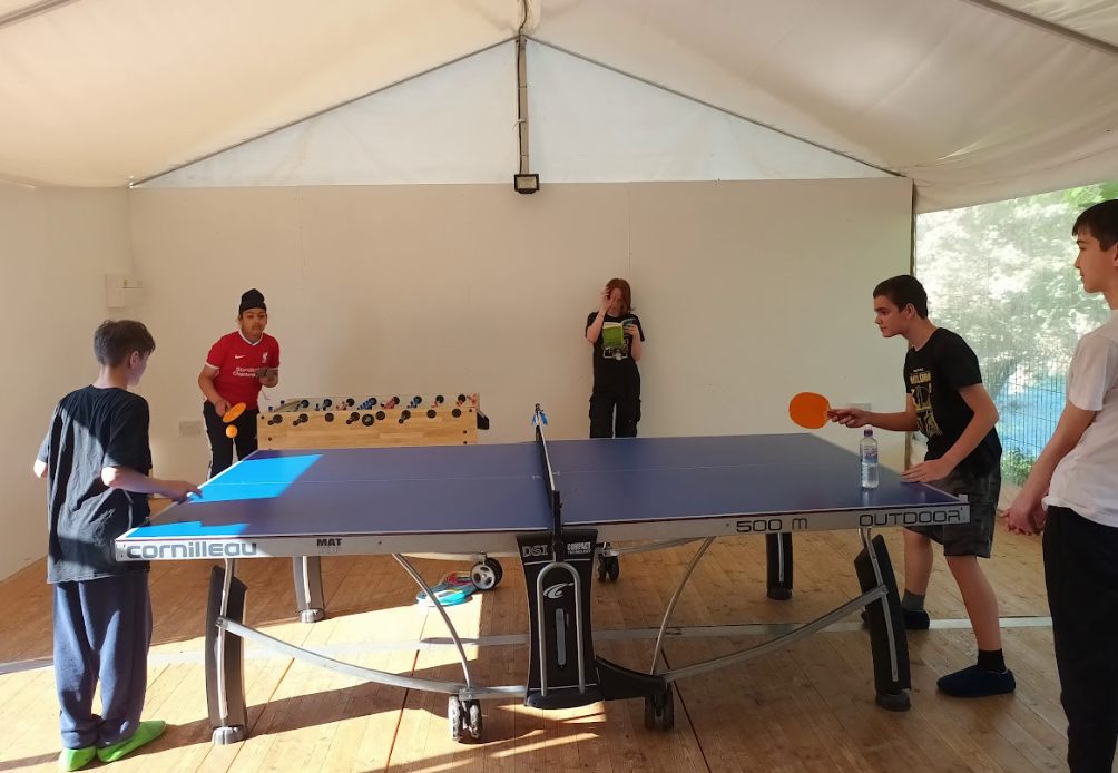The image shows an indoor recreational area, likely a tent or temporary structure, where several young people are engaging in games. In the foreground, there's a blue table tennis (ping-pong) table, with two boys holding paddles, seemingly mid-game. One boy is on the left side of the table, and another is on the right. In the background, to the left, two more individuals are standing near a foosball table, with one of them looking at the camera. Another person is visible further back. The structure has white canvas walls and ceiling. The overall atmosphere suggests a casual and fun environment for various indoor games.