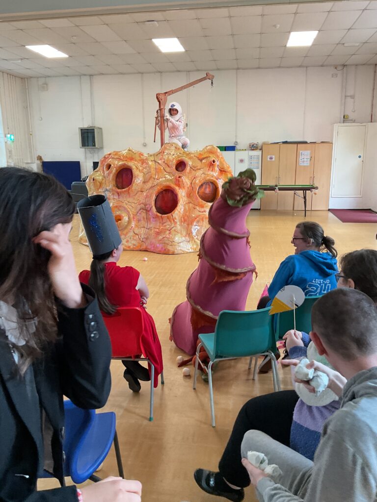 Set in a school hall, there are several people in the foreground and mid-ground, seated on chairs, facing towards a performance area. Some appear to be children or young people, possibly students, and there are also adults present. One person in the foreground is holding what looks like a piece of white clay or dough, suggesting an interactive or workshop element might be part of the event. In the center background, there's a large, elaborate prop that resembles a lunar surface or a large, textured rock formation with multiple holes or craters. Standing on top of this prop is a person in a white or light-colored suit, possibly a costume representing an astronaut or a character related to space. To the right of this large prop, there's another person in a distinctive, colorful costume that is tall and elongated, possibly representing an alien or an abstract creature.