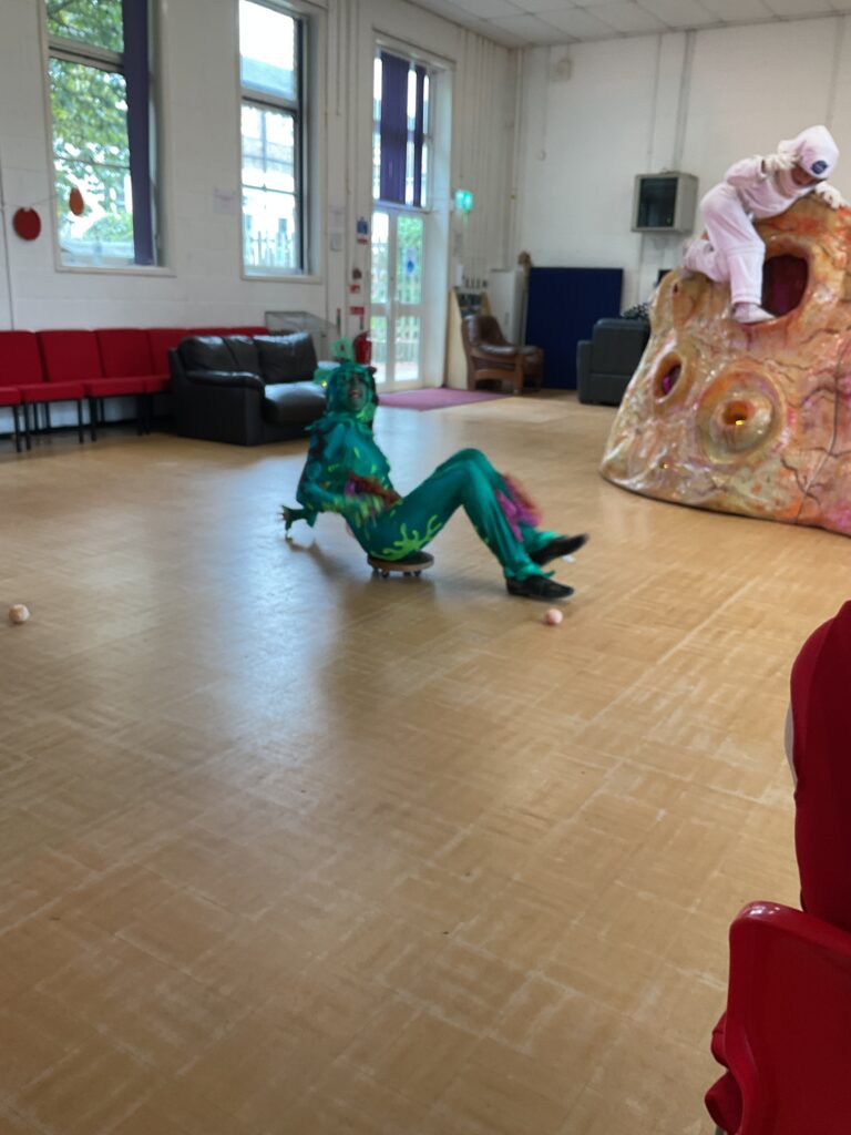 A person in a vibrant green, possibly reptilian or alien-like costume, is positioned on a skateboard or similar low-to-the-ground wheeled device. They are leaning back, creating a dynamic pose that suggests movement or a playful interaction. The costume has various textures and possibly iridescent elements, and the character appears to have horns or elaborate headwear. This is likely the "elongated creature" previously seen in a different pose.