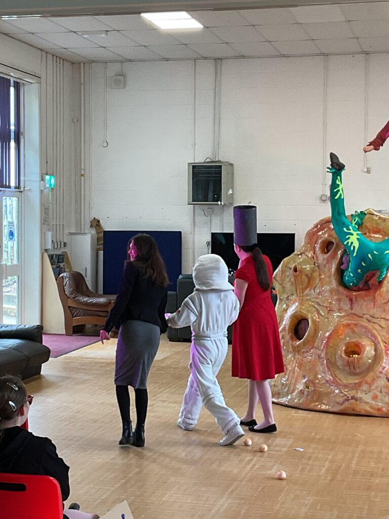 In the mid-ground, there are three individuals with their backs to the camera, walking away from the main performance area towards the left side of the room. The person in the middle is wearing the white astronaut-like suit. To their right, another person is dressed in a red dress with a tall, black, cylindrical hat. This could be another performer or a participant in the event. To the left of the astronaut, an adult, likely a staff member or organizer, is dressed in a black blazer and grey skirt. Their pose suggests they might be guiding the costumed individuals.