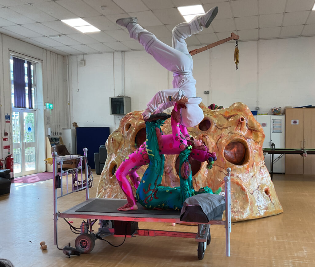 The central focus is a complex acrobatic pose involving two performers. One performer, dressed in the green and pink creature costume (previously identified as the "elongated creature" and the "green creature"), is lying on their back on a mobile platform or bed-like prop. The other performer, dressed in the white astronaut-like suit, is balancing on their hands on the feet of the green/pink performer, performing a handstand or a similar inverted pose. Their legs are extended upwards, and their helmet is off or facing away, revealing the person's head partially.