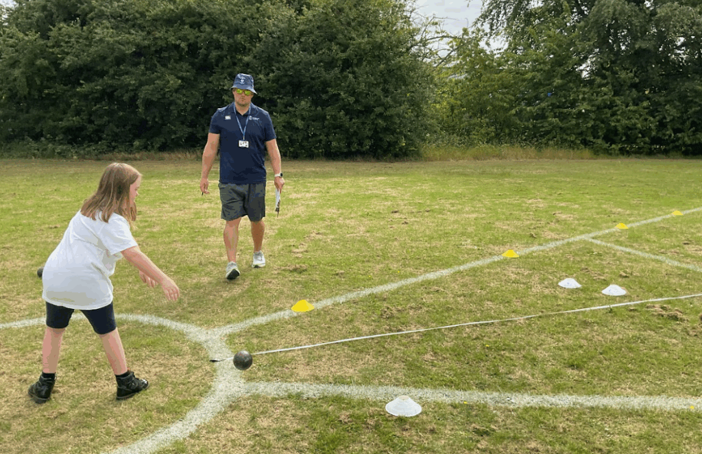 A young pupil taking part in the shot put at Sports day. Watched by a teacher