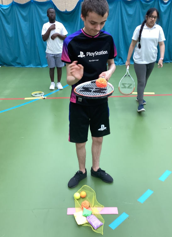 Three young people are on a green sports court with various lines. The person in the center is holding a tennis racket with an orange, textured ball on it, and there are other sports equipment like a tennis racket and small balls on the floor. This seems to be a general sports or skill-building activity, possibly related to racket sports or hand-eye coordination drills.