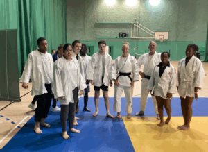A group of people, both young and adult, are standing on a blue and yellow mat in what looks like a martial arts dojo or training area. Most of them are wearing white martial arts gi, with one woman wearing a black belt, indicating a higher rank. This image suggests a judo or similar martial arts session.
