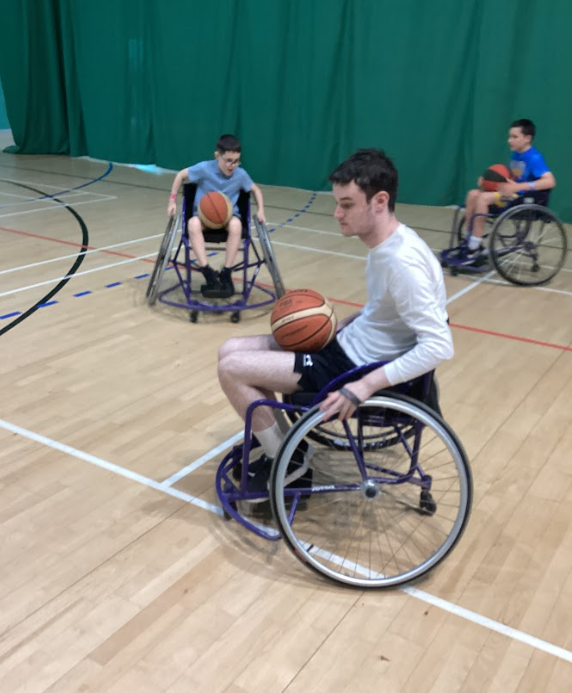 Three young people in wheelchairs are on a basketball court, each with a basketball. This clearly shows a wheelchair basketball activity or practice sessio