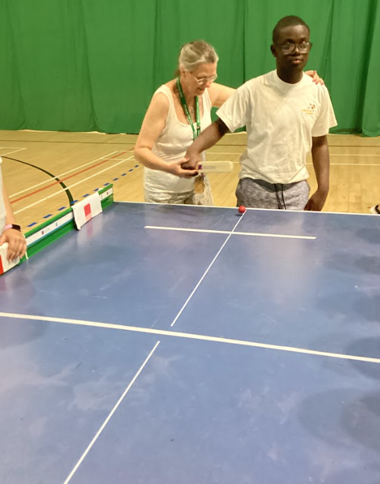 This image appears to show two people, an adult woman and a young person, engaged in an activity that looks like a modified form of table tennis or a similar tabletop game. The young person, wearing glasses and a white t-shirt, is positioned at a blue table with white lines, which resembles a ping-pong table. Their right arm is extended towards the table, and they are looking towards the right side of the frame. An adult woman, wearing a light-colored top and what looks like a lanyard, is standing behind and slightly to the left of the young person, with her hand on their shoulder. She seems to be offering guidance or support. There's a red ball visible on the table, and to the left, there's a green and white structure that might be part of the game setup, possibly a ramp or a guide for the ball. Another person's arm is partially visible on the far left, suggesting more participants. The setting appears to be a gymnasium or a large indoor space with a green wall in the background.