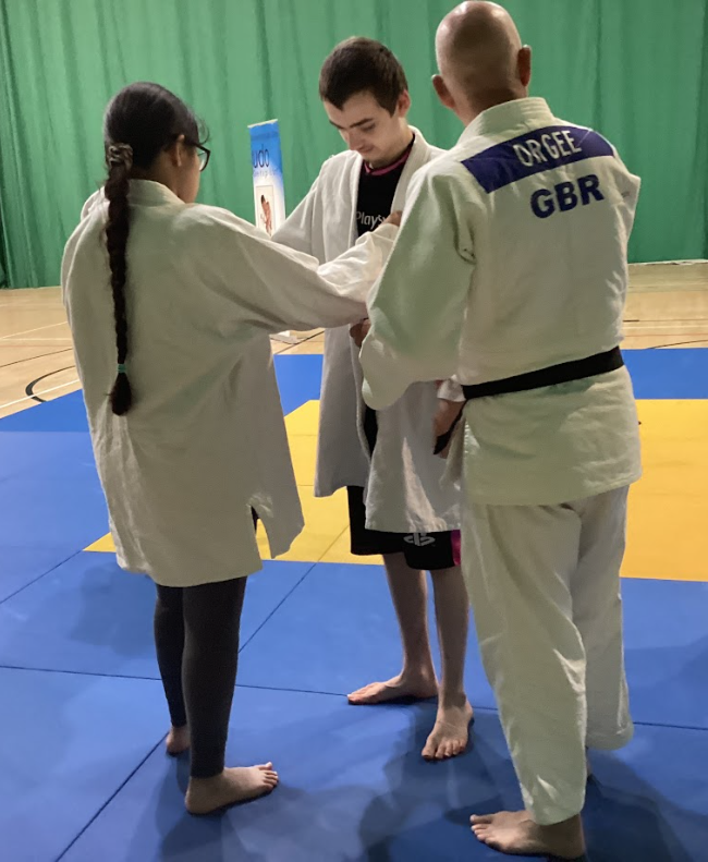 This picture shows three individuals on a blue and yellow mat, which is characteristic of a judo or martial arts dojo. The person on the right is an adult male wearing a white judo gi with "ORGEE GBR" on the back, suggesting a coach or instructor. He is facing a young man who is also wearing a gi (or partially wearing one). A young woman with a long braid is standing to the left, also in a gi, and appears to be receiving instruction or getting ready to practice with the young man. This looks like a judo training session.