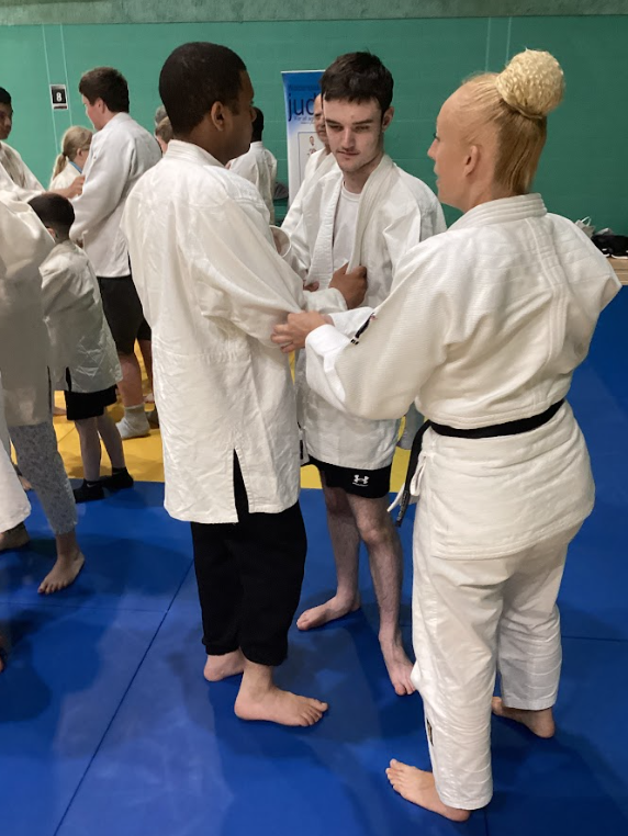 this image takes place on a judo mat with multiple participants in white judo gi. In the foreground, an adult woman with blonde hair tied in a bun, wearing a gi with a black belt (indicating a high rank, likely an instructor), is instructing or demonstrating something to two young men. All three are facing each other, and the instructor's hands are on the gi of one of the young men, demonstrating a grip or technique. Other participants are visible in the background, also in gi. This confirms a judo training session is in progress, with instruction being given.