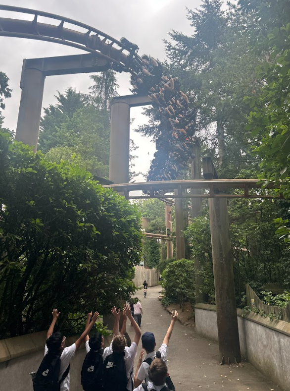 A group of children are looking up and waving their hands at a large roller coaster with people on it, which is suspended high above them on the track. This shows excitement and interaction with a major park attraction.
