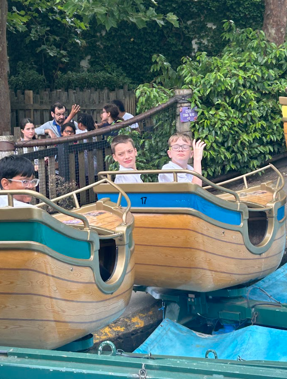 Children are riding in small, wooden boat-like carriages on a water ride. Some are smiling and waving.