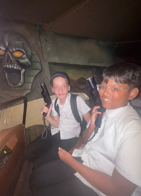 Two boys are inside what looks like a dark ride or interactive attraction, holding laser guns and aiming them. A skull-like image is visible on the wall behind them.