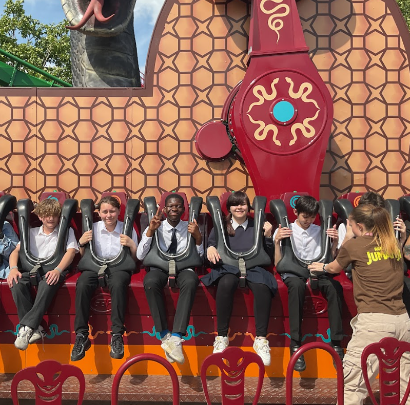 A group of children are seated on a ride, strapped in, and smiling. A staff member is assisting them. This is clearly a theme park ride.