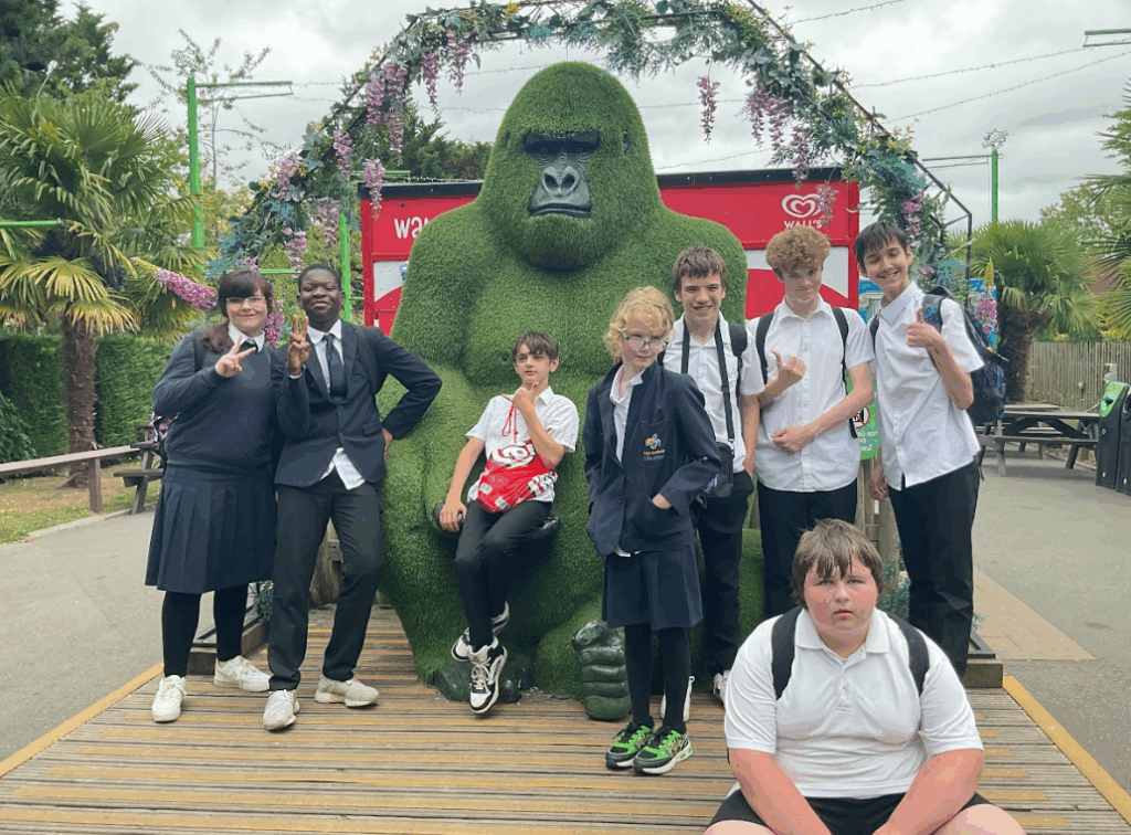 A group of children and young people, mostly in white shirts and dark trousers/skirts, are posing for a photo in front of a large, green gorilla statue. There are palm trees and other park elements in the background. This is another scene from a leisure park.