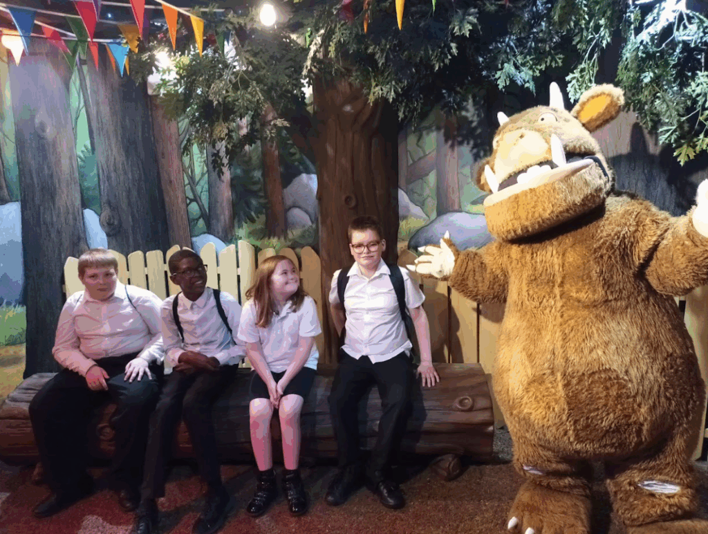 Four children (three boys and one girl, all still in their school-like attire) are sitting on a wooden log in a themed area that resembles a forest. They are posing with a large mascot character that looks like the "Gruffalo," a popular children's book character. The background has colorful bunting, indicating a festive or dedicated themed zone within the park.