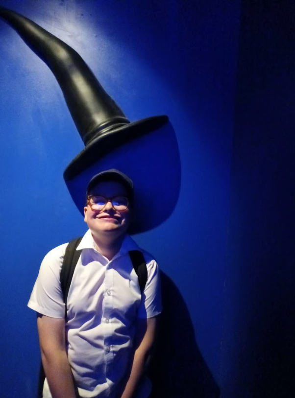 A person in a white shirt and glasses is smiling, standing in front of a blue wall with a large, dark witch's hat structure above them, creating a shadow on the wall. This looks like an indoor exhibit or photo opportunity at an attraction.
