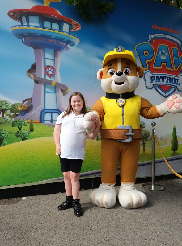 A young girl is standing next to a large mascot character of Rubble from Paw Patrol, with a Paw Patrol themed backdrop behind them. This confirms a theme park or children's attraction.