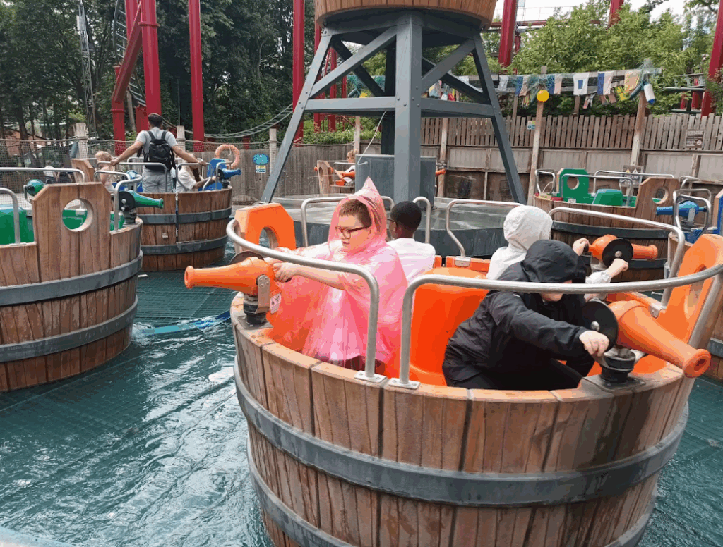 Children are in barrel-shaped boats on a water ride, some wearing ponchos. They appear to be operating water cannons. This is another theme park water ride.