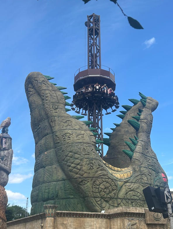 A tall drop tower ride is seen against a blue sky, with riders at the top. The base of the tower is elaborately designed to look like the open jaws of a large, monstrous creature (possibly a crocodile or alligator), adding to the theme park's immersive design.