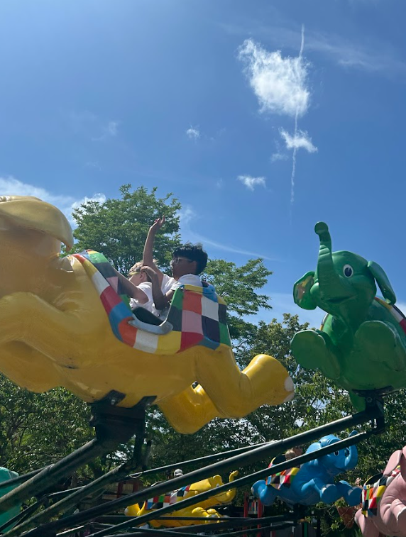 Children are on an aerial carousel ride featuring colorful elephant-shaped carriages, against a bright blue sky. One child is waving.