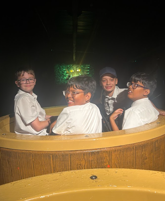 Four boys are sitting in a large, barrel-shaped boat, possibly an indoor water ride or dark ride, with a cartoonish, green, glowing face in the background. They are smiling and looking at the camera.