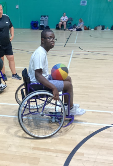 A young person in a wheelchair is sitting on a basketball court, holding a multicolored basketball. Another adult is partially visible to the left. This suggests participation in wheelchair sports.