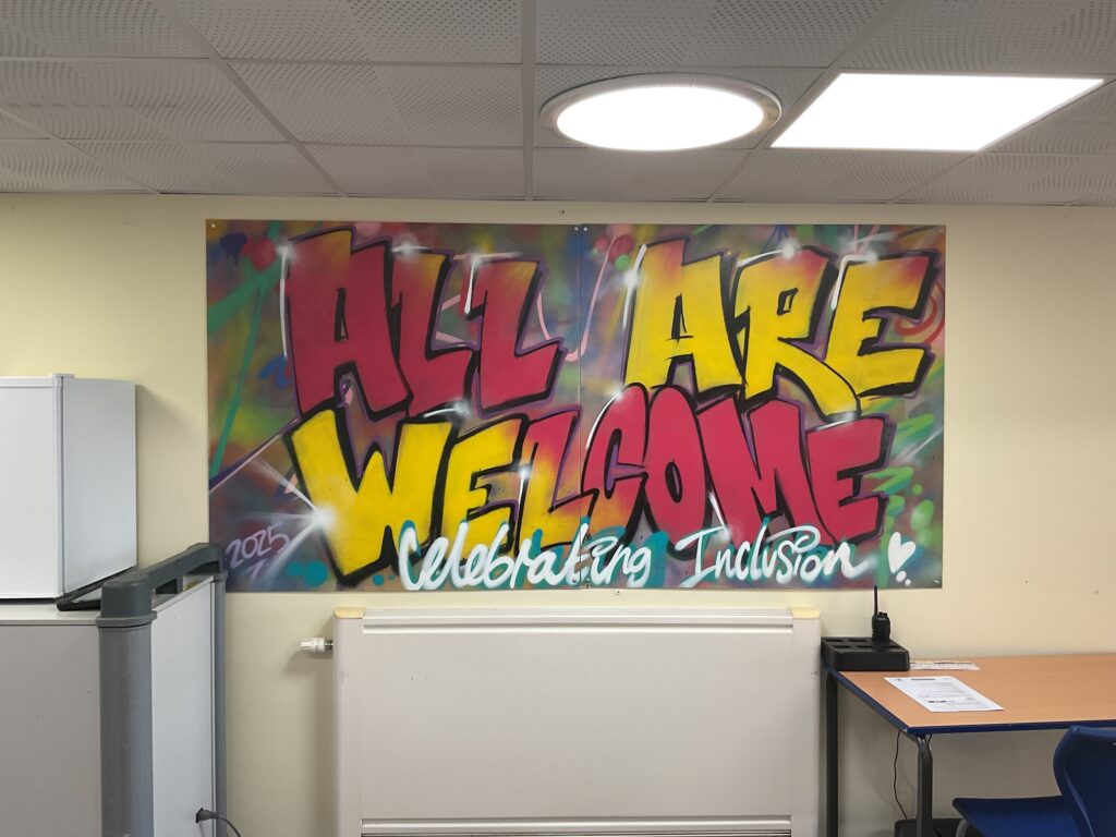An indoor image featuring a large piece of graffiti art on a wall. The art displays the words "ALL ARE WELCOME" in large, bold red and yellow letters, with smaller writing underneath that says "Celebrating Inclusion." The room has a white ceiling and yellow walls.