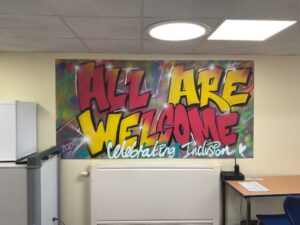 An indoor image featuring a large piece of graffiti art on a wall. The art displays the words "ALL ARE WELCOME" in large, bold red and yellow letters, with smaller writing underneath that says "Celebrating Inclusion." The room has a white ceiling and yellow walls.