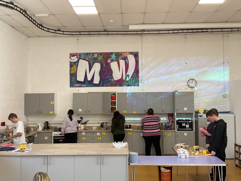 This image shows an indoor setting, likely a cafe or kitchen area, with several young people preparing food or drinks. There are grey cabinets and appliances, and a large piece of colorful graffiti art spelling out "M.V. cafe" is displayed on the wall above the cabinets.